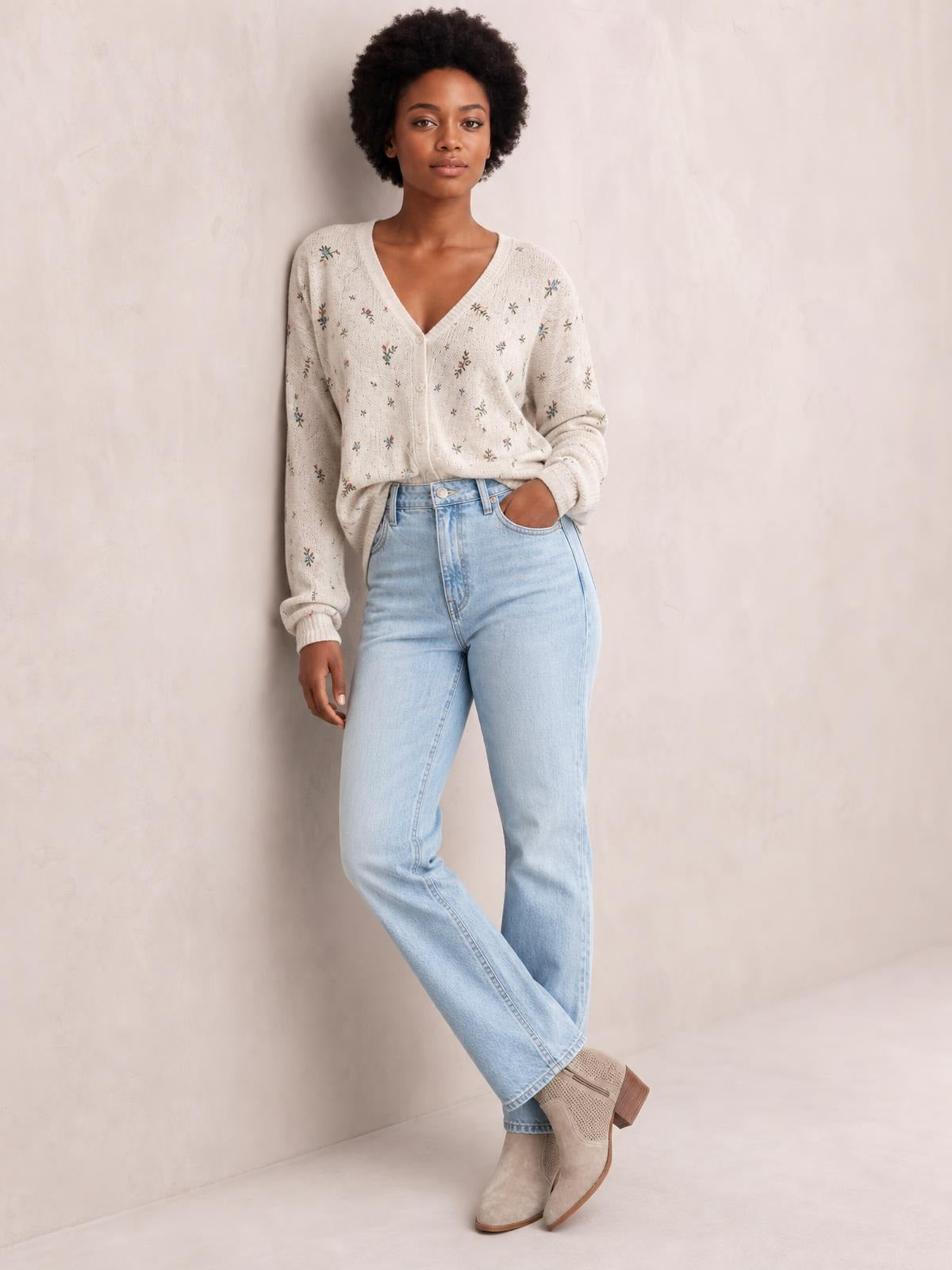 A model poses in a cream floral V-neck button-down cardigan with long sleeves, ribbed cuffs, and a relaxed silhouette, paired with light-wash jeans.