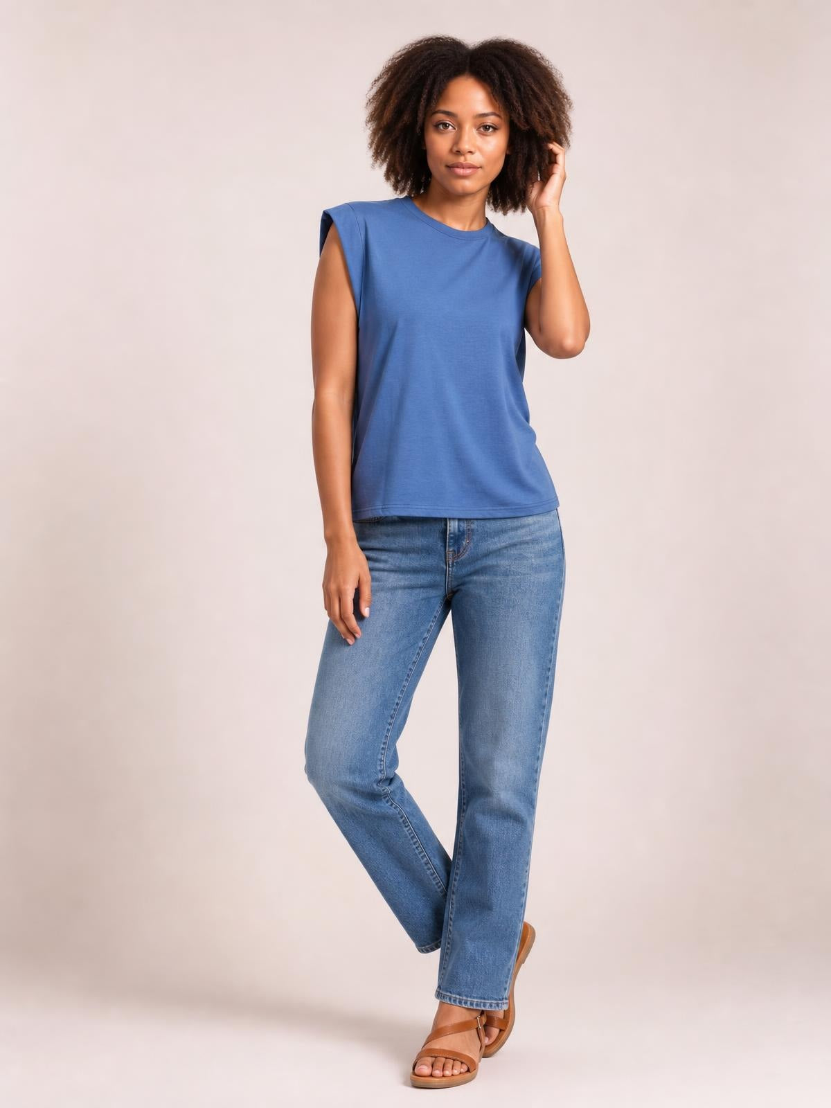 A model wears a blue Florira round neck cap sleeve T-shirt with a relaxed straight silhouette, posing with one hand in her hair and crossed legs.