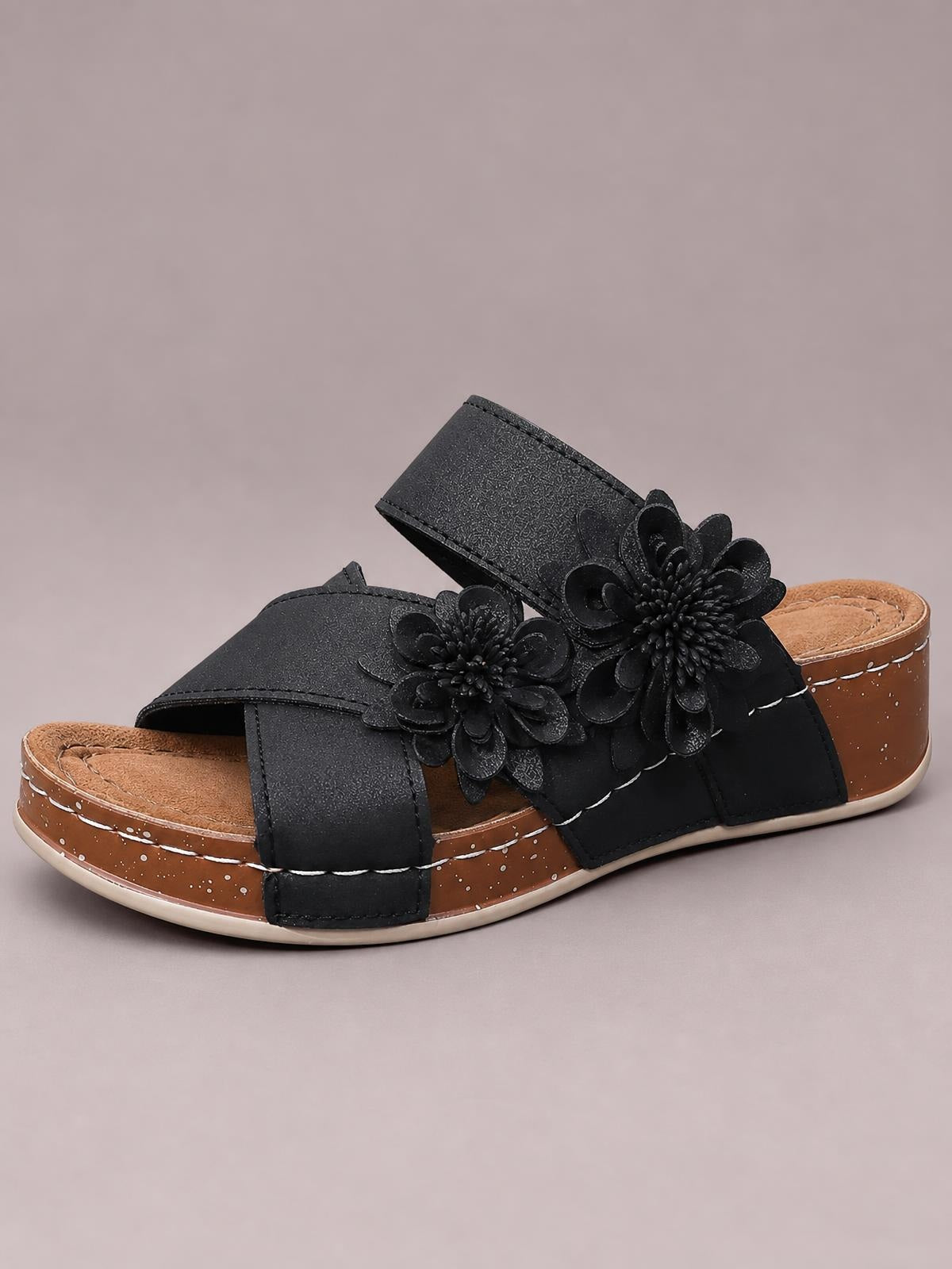 Black flower crisscross wedge sandals feature layered floral embellishments, wide stitched straps, open toes, and a mid-height brown wedge sole.
