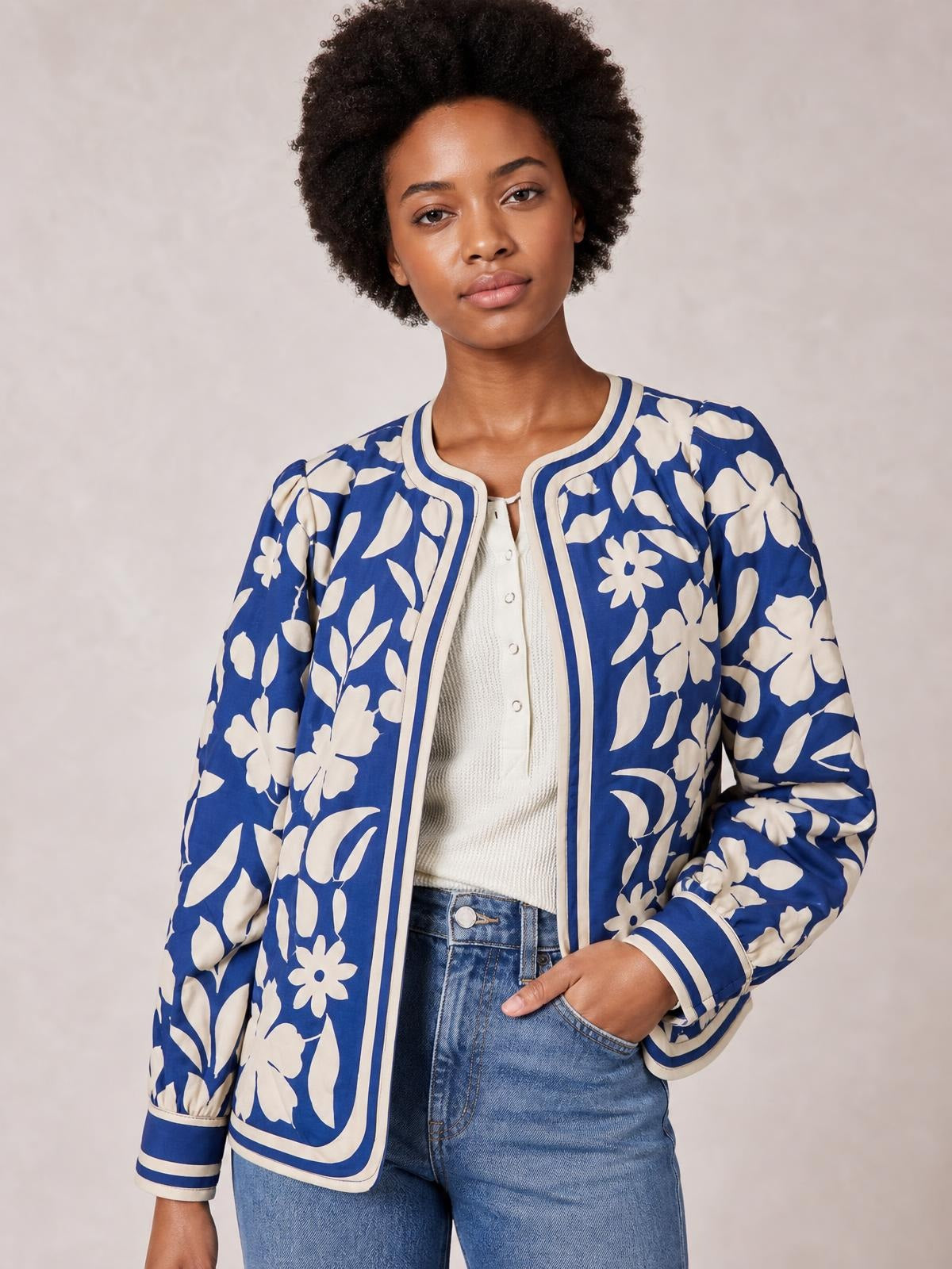 A model wears a blue and cream flower print jacket with an open front, long sleeves, contrast trim, and buttoned cuffs, posing with one hand in a pocket.
