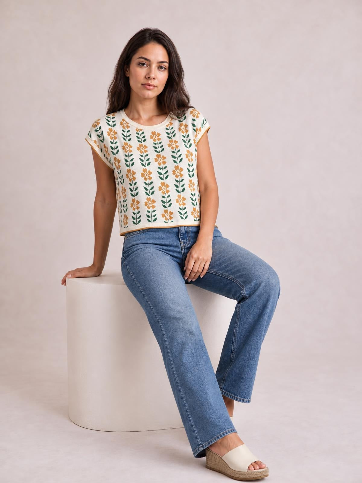 A woman sits in blue jeans wearing a cream floral knit top with a round neck, short sleeves, relaxed cropped silhouette, and tan-trimmed edges.