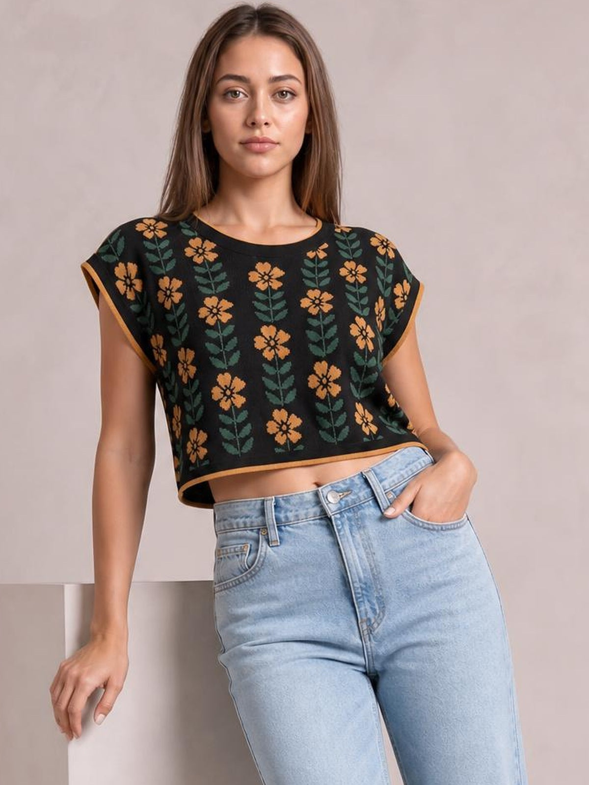 Model wearing a black floral knit top with a round neck, short sleeves, orange trim, and a cropped, relaxed silhouette while posing with one hand in her pocket.