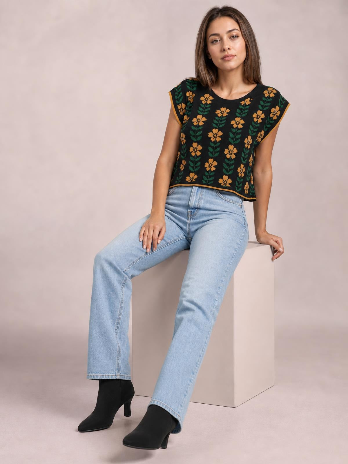 A seated model wears a black flower knit top with orange floral and green leaf motifs, a round neck, short sleeves, and a relaxed straight silhouette.