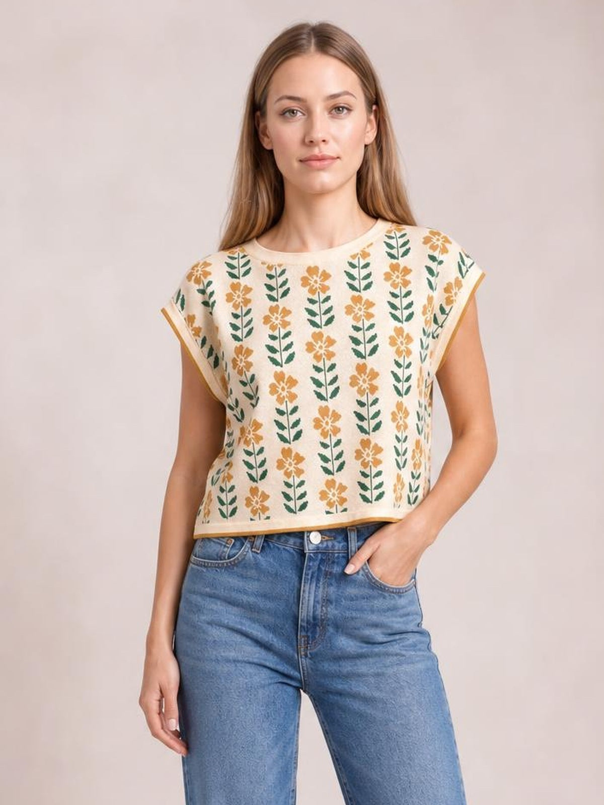 A model wears a cream knit top with a round neck, short sleeves, green-and-gold floral print, contrast trim, and a relaxed cropped silhouette, posing with one hand in her jeans pocket.