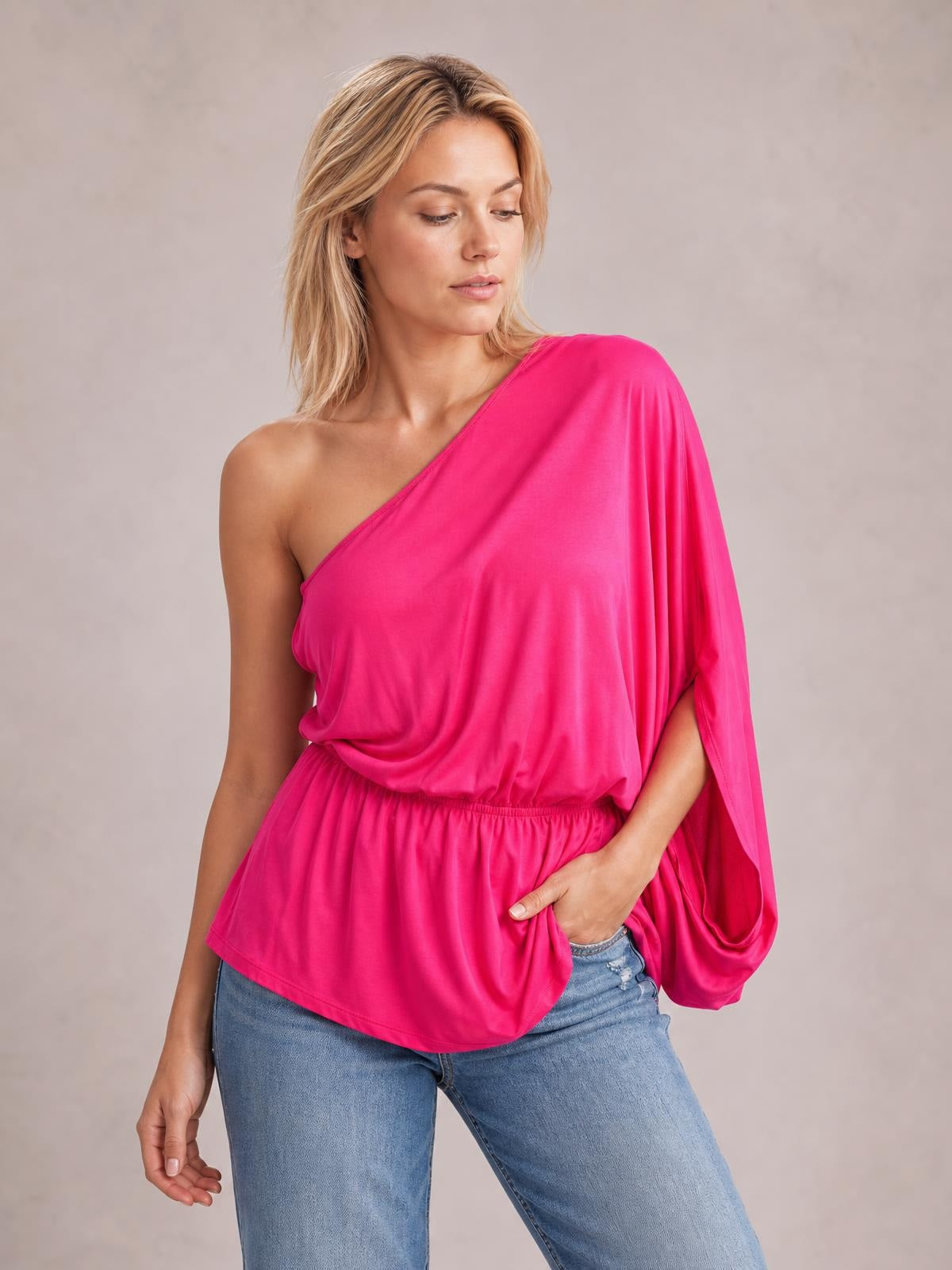 Model poses in a bright pink one-shoulder top with an asymmetrical neckline, single long sleeve, relaxed draped silhouette, and elastic waist.