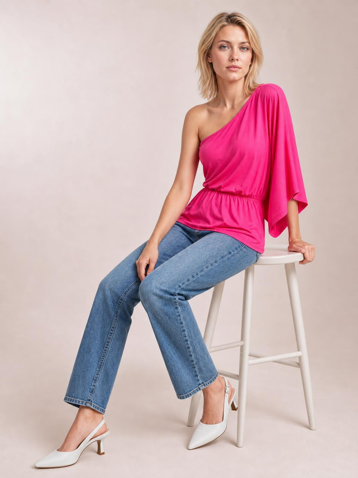 Model seated on a stool wears the bright pink Forever And Always one-shoulder top with an asymmetrical neckline, long single sleeve, and elasticized waist.