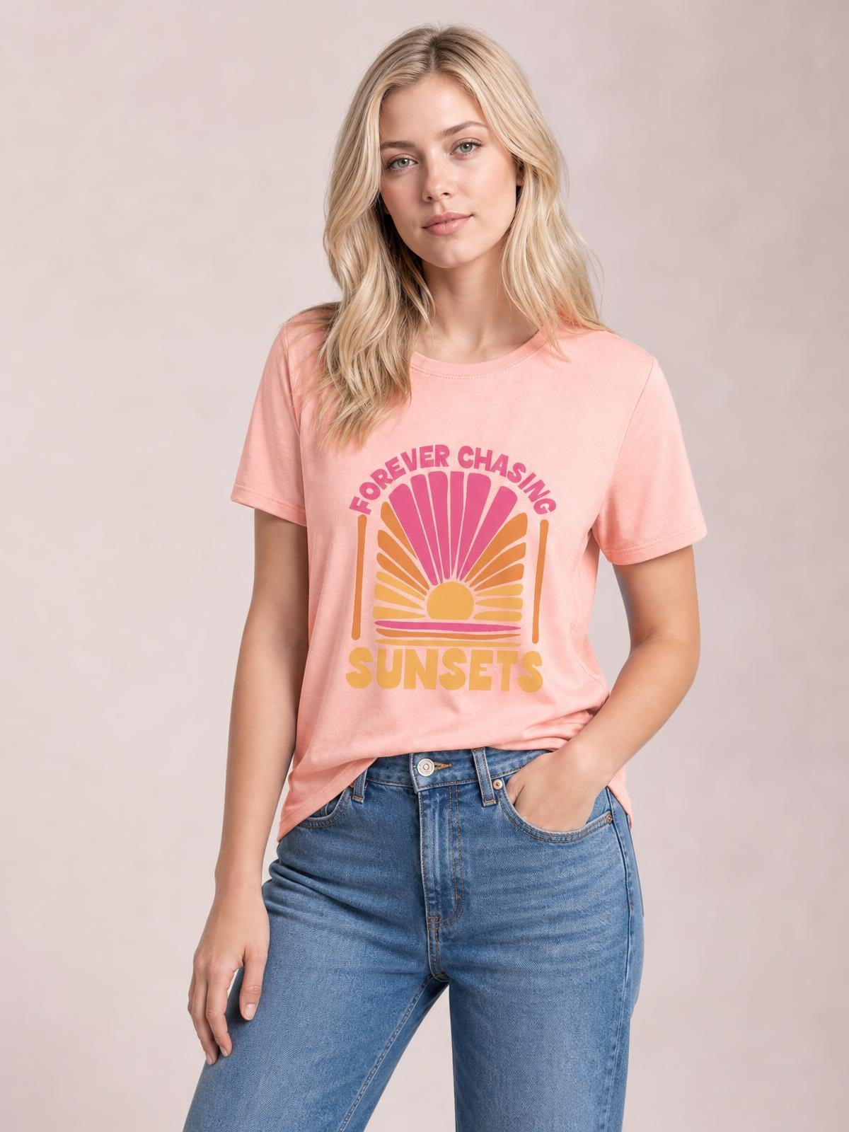 A model wears a pink round neck T-shirt with a bold “FOREVER CHASING SUNSETS” graphic, short sleeves, and a relaxed silhouette, posing with one hand in her jeans pocket.