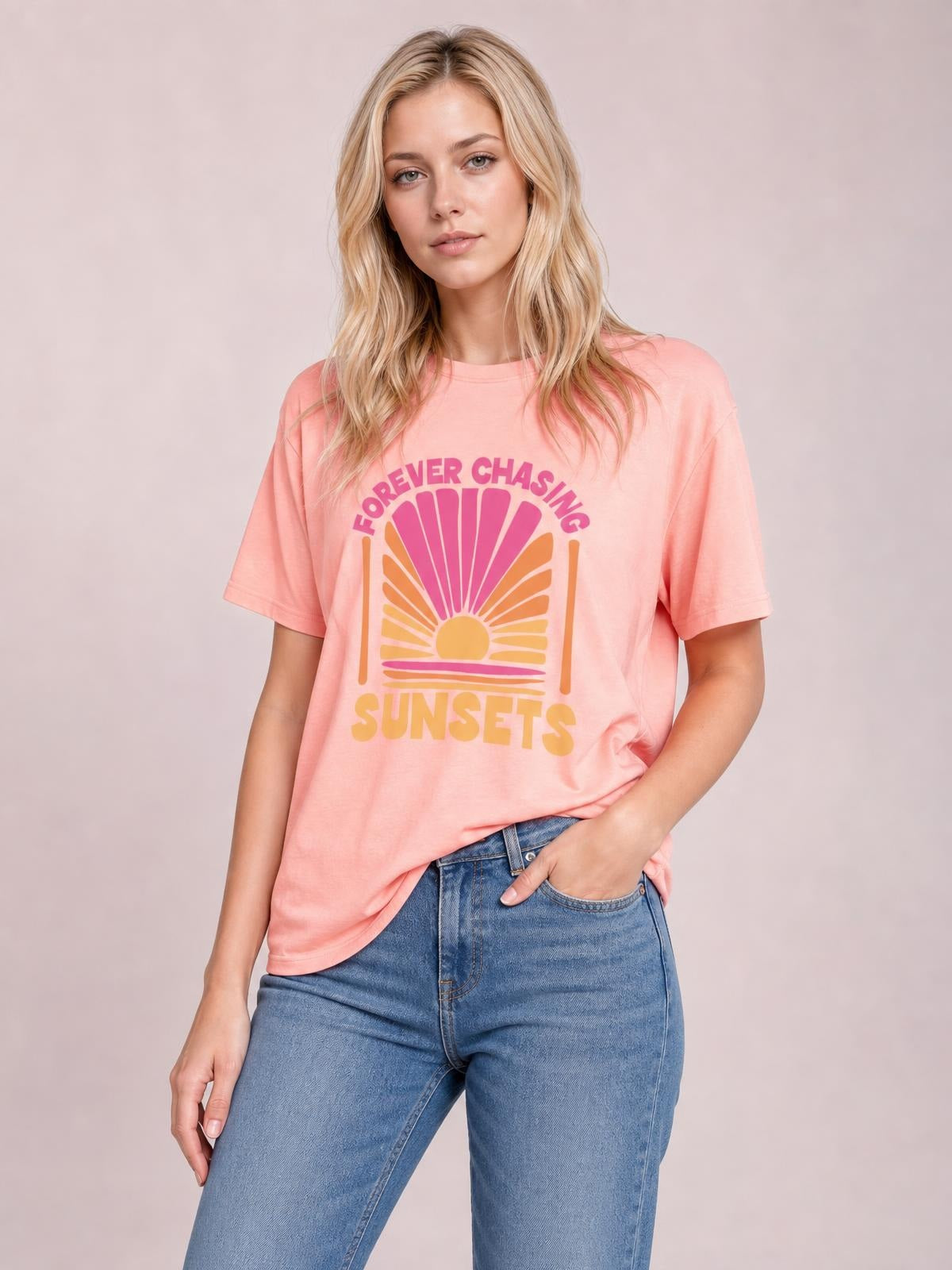 Model wears a relaxed pink round neck T-shirt with short sleeves, a sunset graphic, and “FOREVER CHASING SUNSETS” lettering, paired with blue jeans.