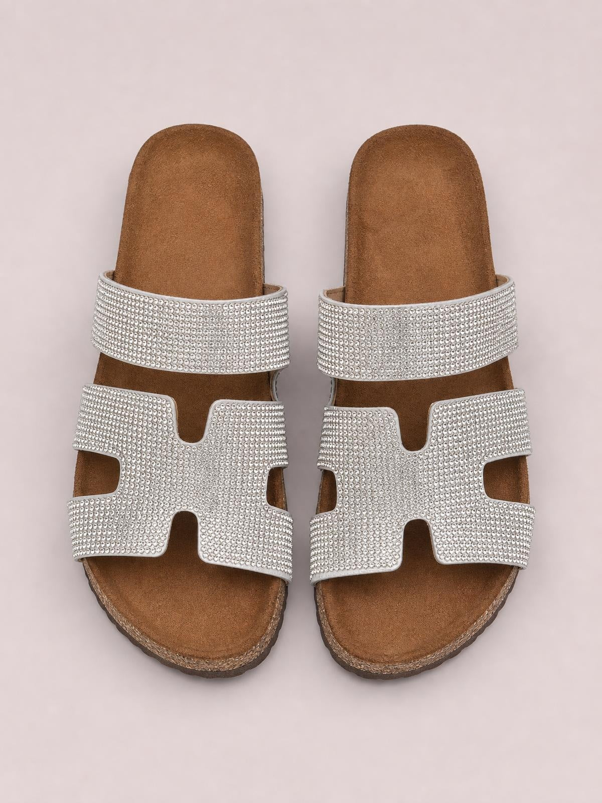 Silver rhinestone open toe sandals with wide embellished straps, a cushioned brown footbed, flat sole, and structured cutout construction.