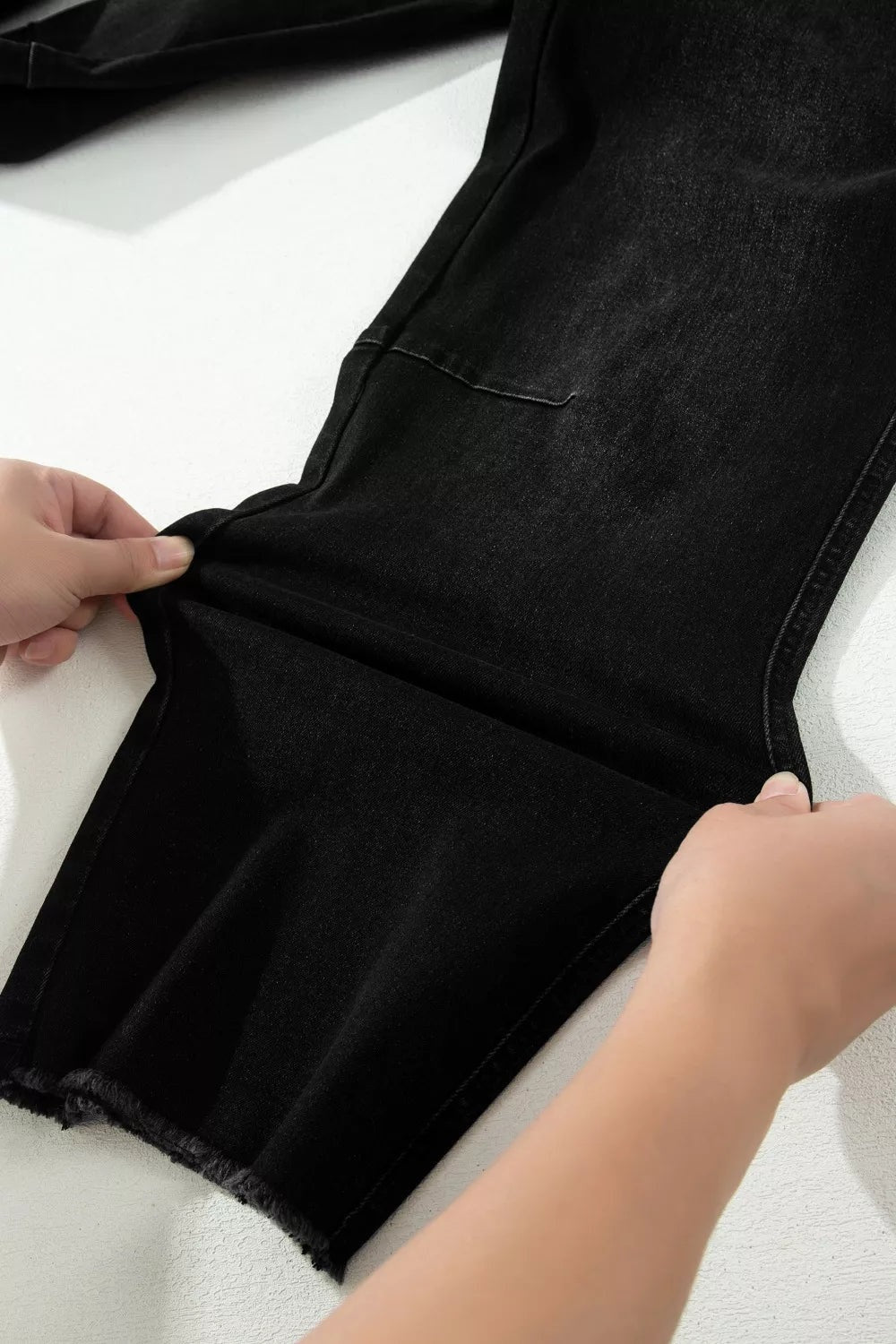 Close-up of black frayed cropped straight jeans showing the pocketed leg, visible seams, and raw hem as hands hold the fabric flat.