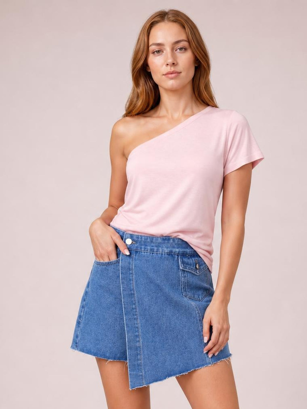Model stands with one hand in a pocket, wearing a blue denim skort with an asymmetrical silhouette, front seam, patch pockets, button closure, and frayed hem.