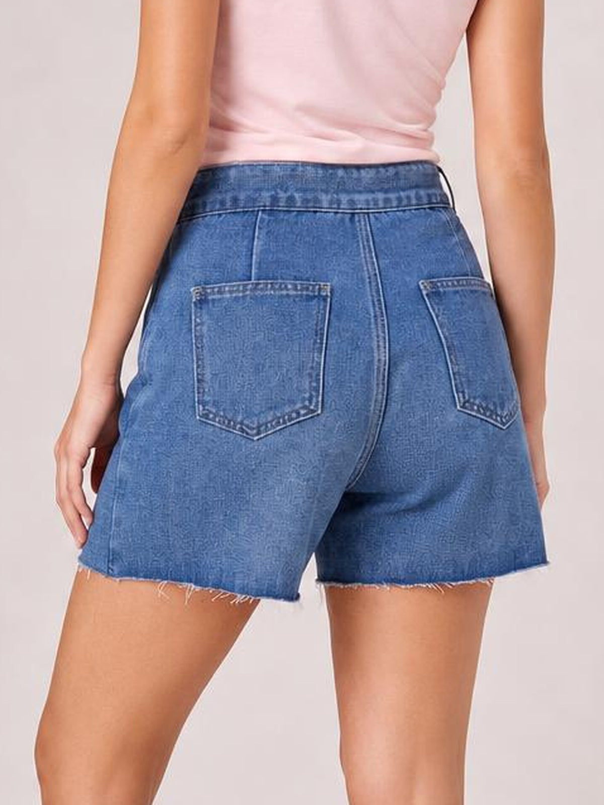 Model shown from behind wearing a blue denim skort with an asymmetrical frayed hem, structured waistband, back patch pockets, and a relaxed short silhouette.