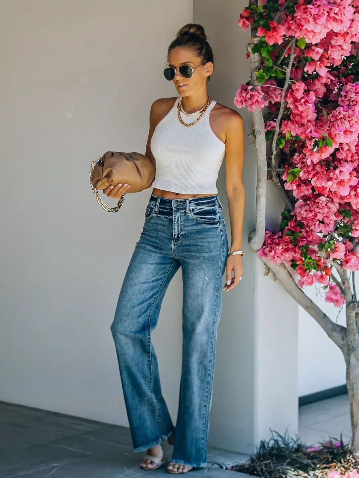 Model stands holding a handbag in blue high-waisted wide-leg jeans with front pockets, contrast fading, and raw frayed hems.