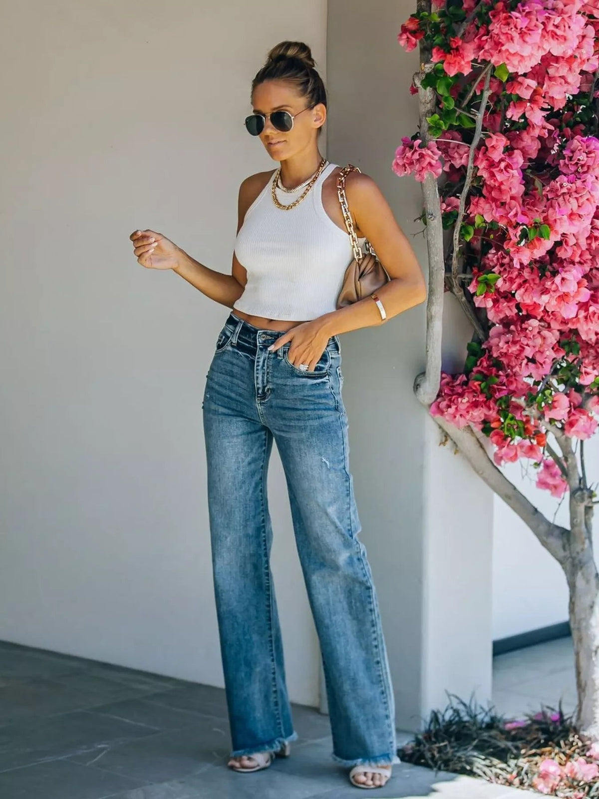 A model stands in blue high-waisted wide-leg jeans with front pockets, belt loops, a button fly, and raw frayed hems, one hand tucked into a pocket.