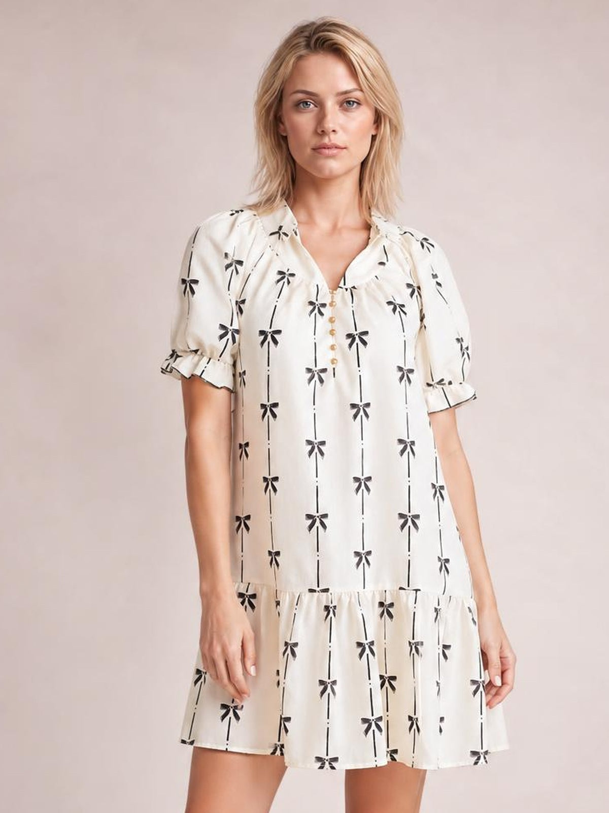 Model wears an opaque white mini dress with black bow print, short frill-trimmed sleeves, a notched neckline, button front, and tiered relaxed silhouette.