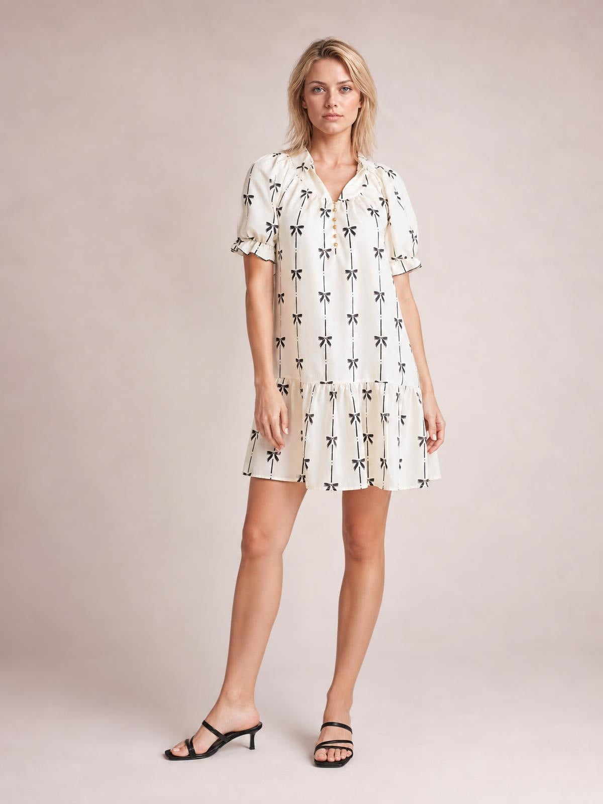 Model stands in a white bow-print mini dress with a notched neckline, short frill-trimmed sleeves, button front, and relaxed tiered silhouette.