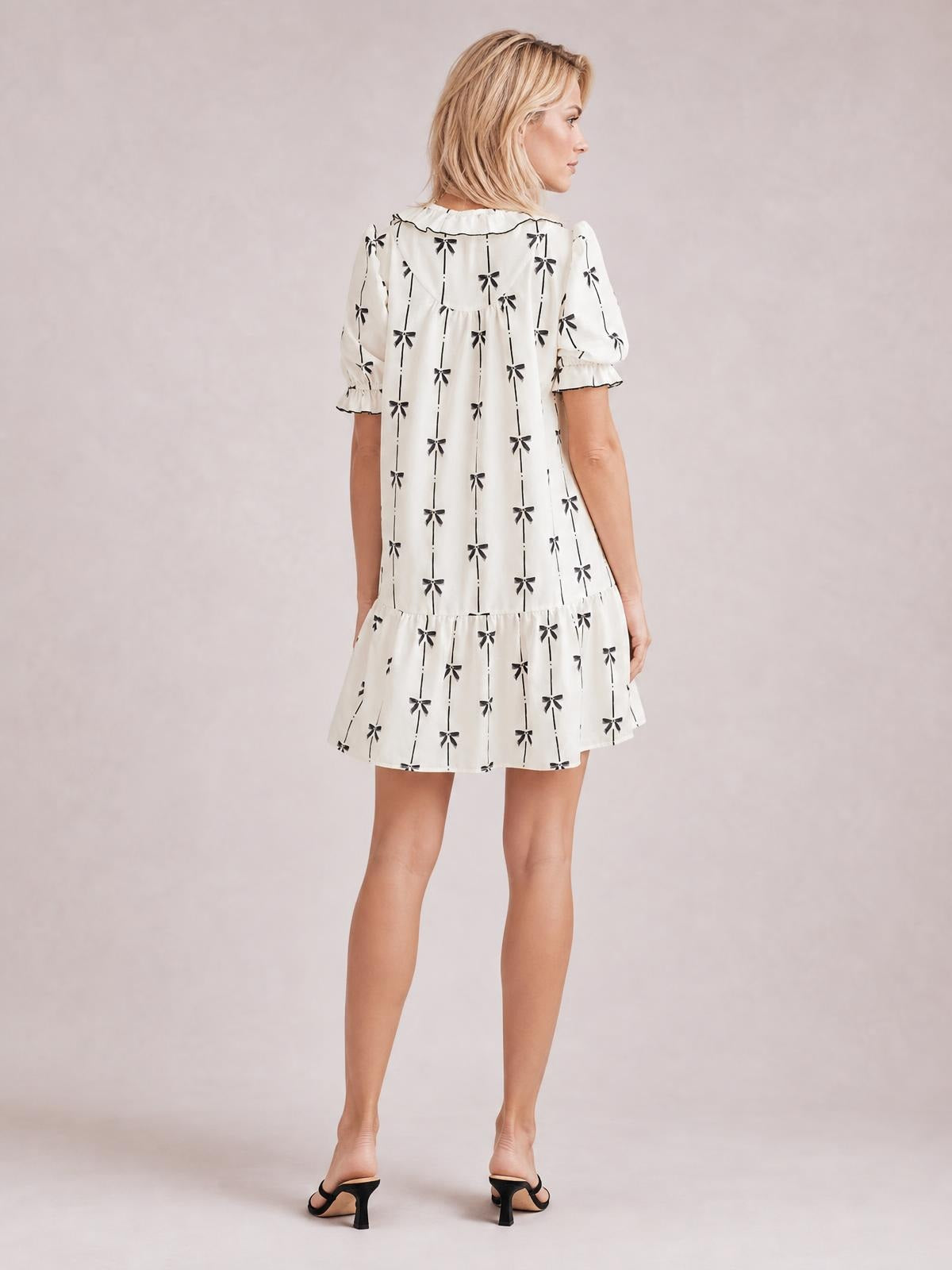 Model turned away showcases an opaque white mini dress with black bow print, short frilled sleeves, a loose silhouette, gathered waist, and ruffled hem.
