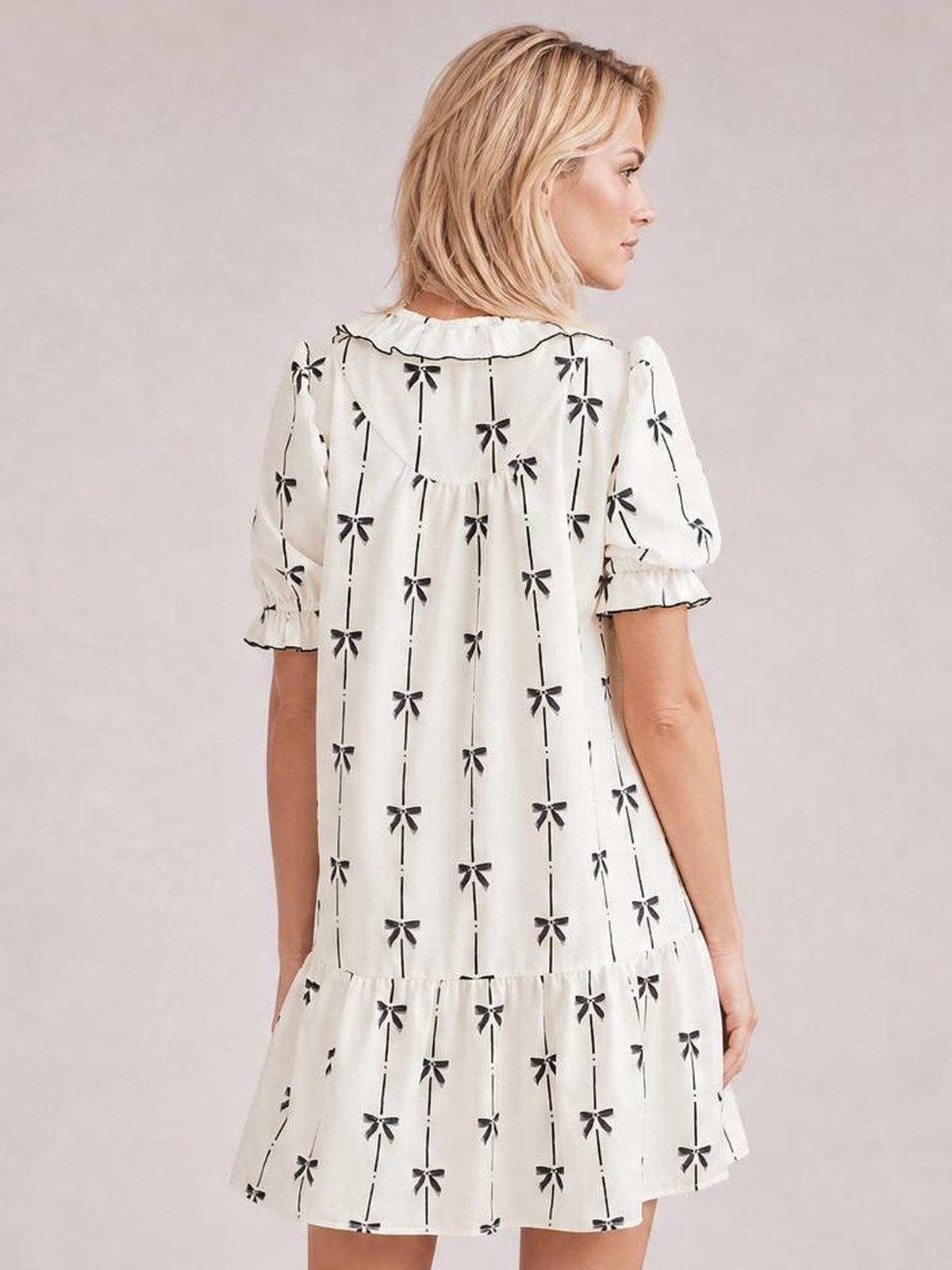 Model shown from behind in a white bow-print mini dress with short frilled sleeves, a loose A-line silhouette, and a gathered tiered hem.