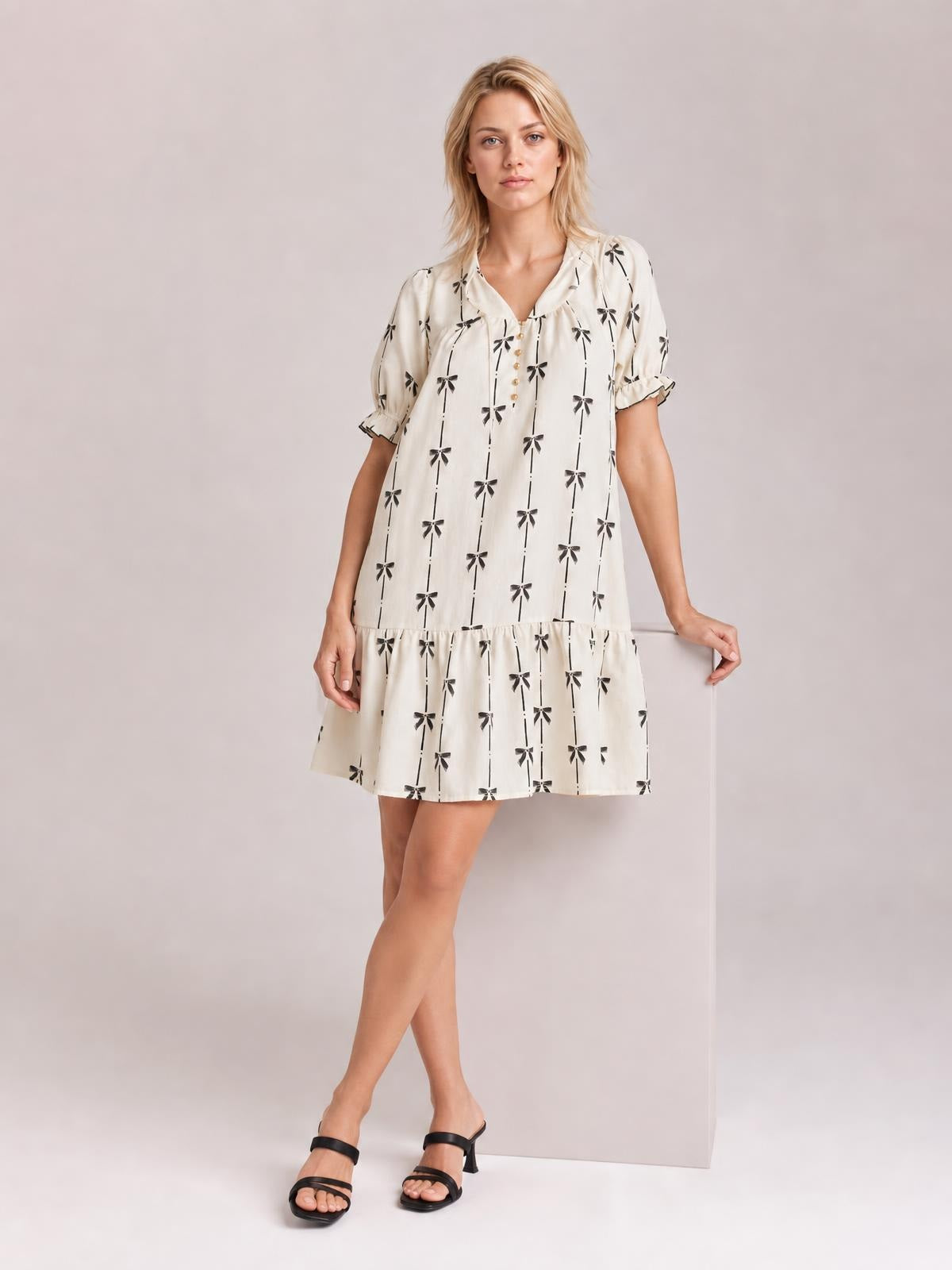 Model wears an ivory mini dress with black bow print, a notched neckline, short frill-trimmed sleeves, and a gathered tiered skirt while standing with crossed legs.