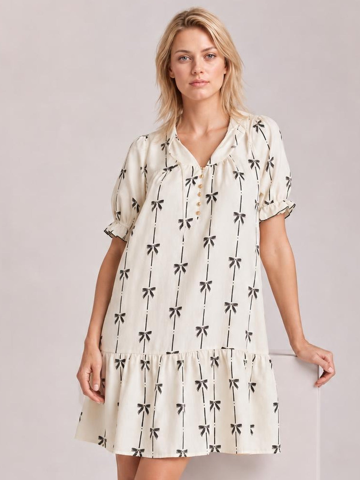 A model wears an ivory mini dress with black bow print, a notched ruffle neckline, short frilled sleeves, and a loose tiered silhouette.