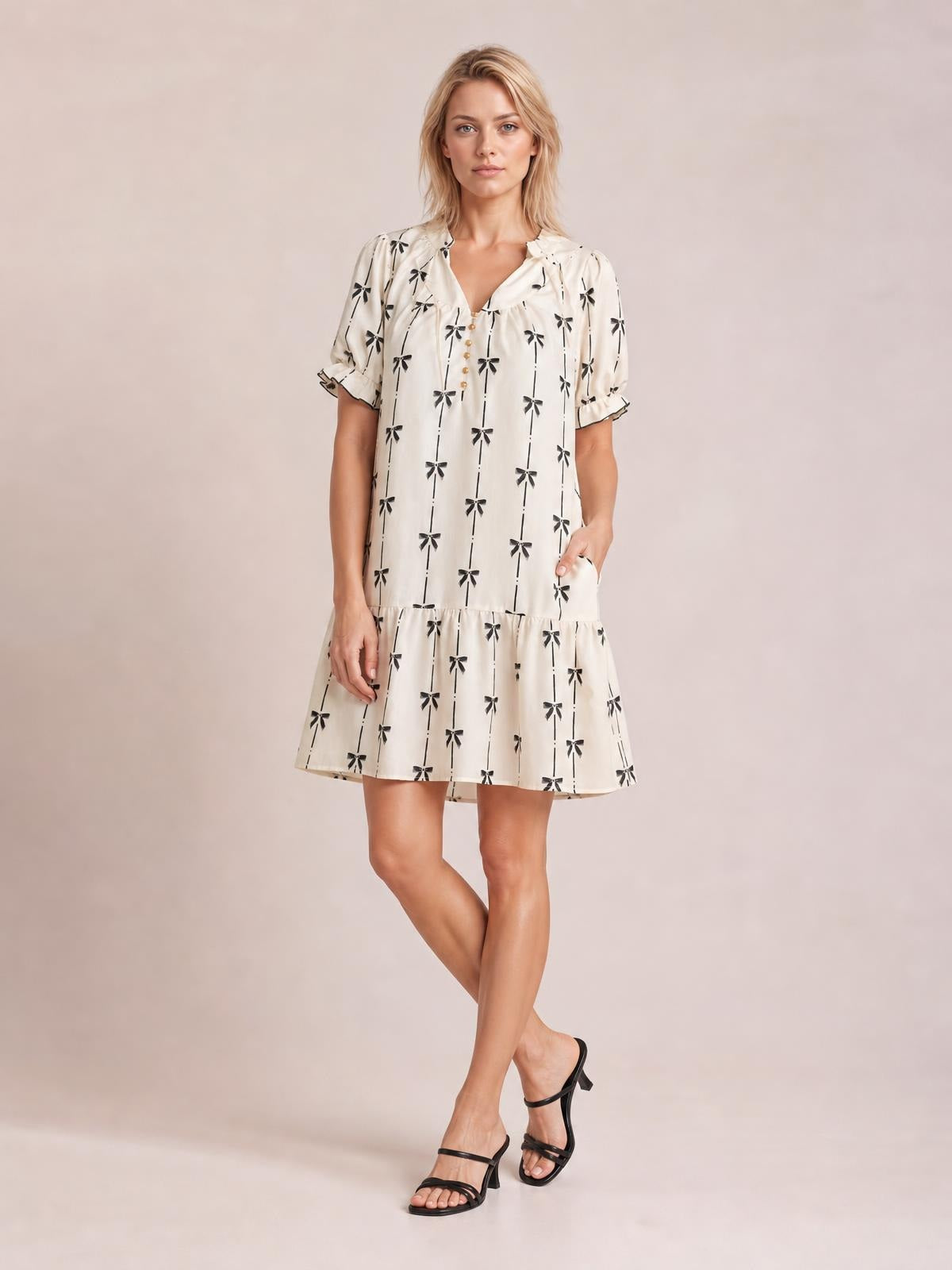 A model stands with crossed legs and one hand in a pocket, wearing an ivory bow print mini dress with a notched neckline, button front, frilled short sleeves, and a flared hem.