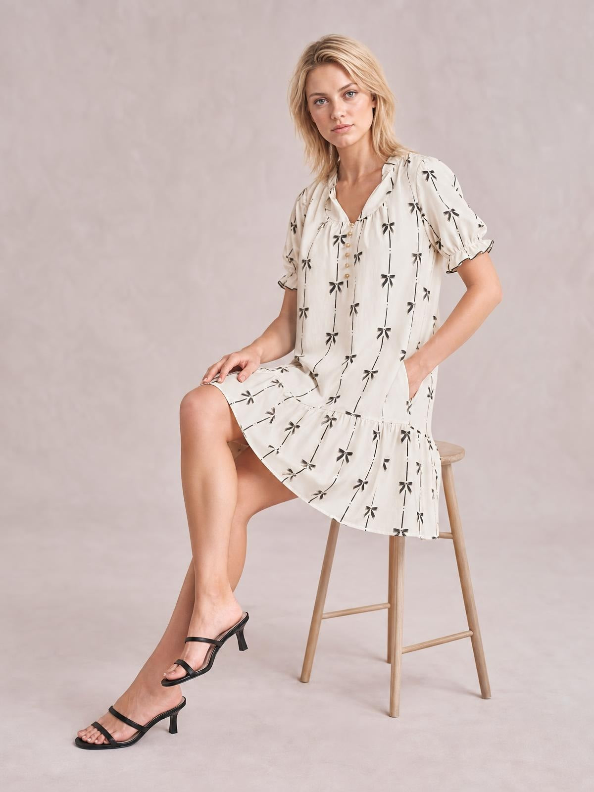 A seated model wears an ivory mini dress with black bow print, a notched neckline, short frilled sleeves, button front, and a relaxed tiered silhouette.