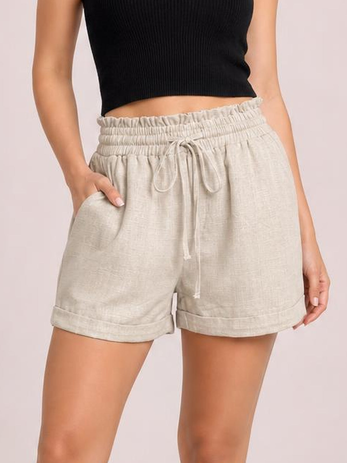 Model wears light beige frill drawstring shorts with a relaxed silhouette, elastic waistband, front pockets, and rolled cuffs, posing with one hand in a pocket.