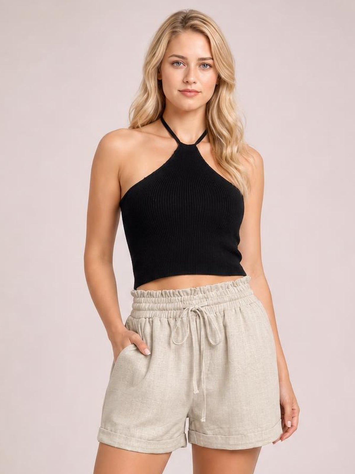 A blonde model wearing a black ribbed halter crop top and light beige drawstring shorts, standing against a solid pale pink studio background.