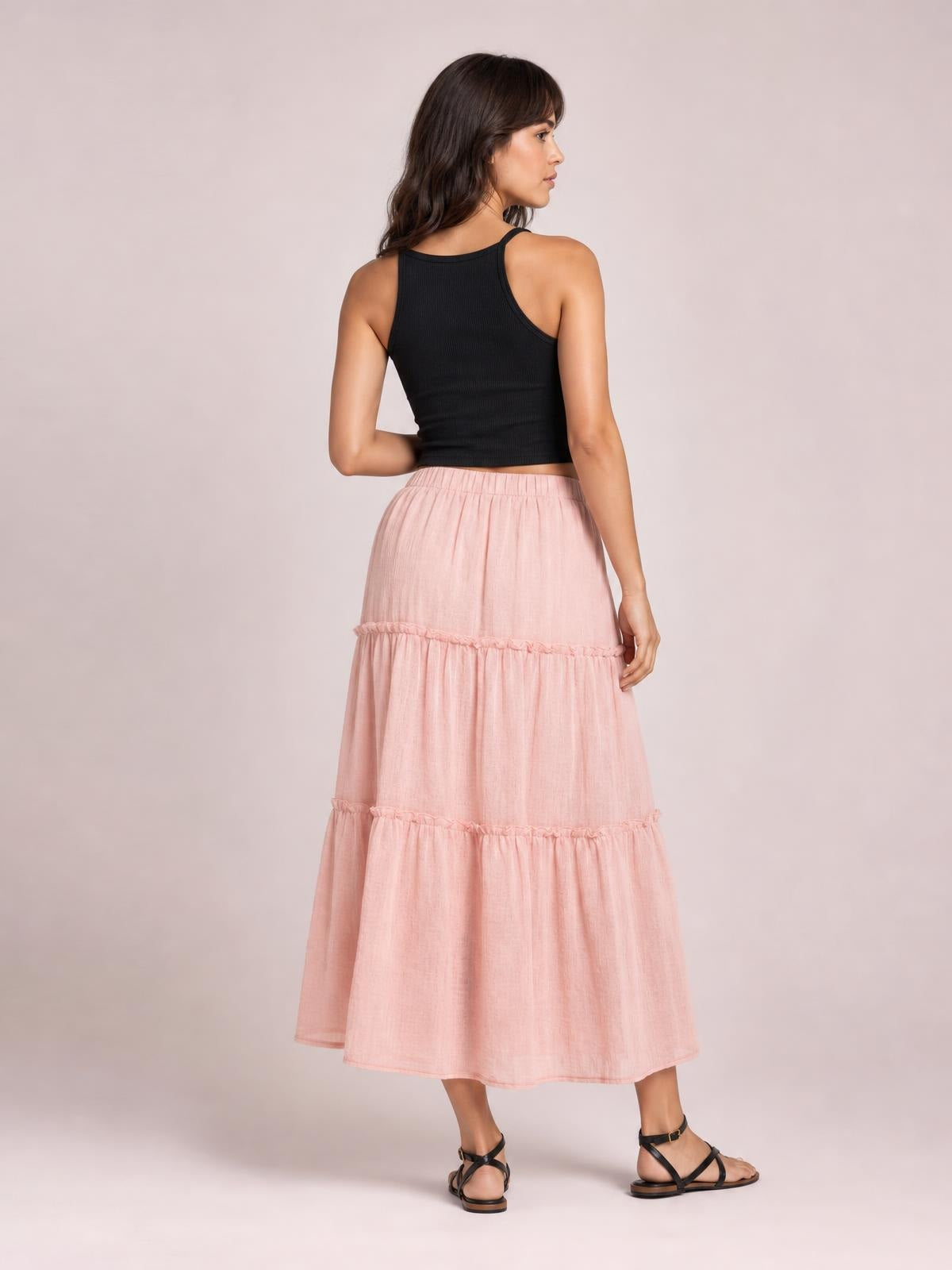Model in a back-facing pose wears a light pink frill elastic waist skirt with tiered ruffles and a flowing midi silhouette.