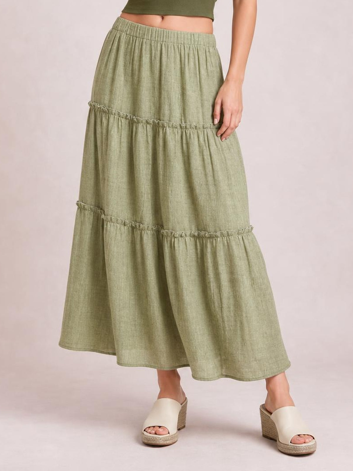 Model wears a sage green Frill Elastic Waist Skirt with a gathered waistband, tiered silhouette, and two ruffled seams, styled at a flowing midi length.