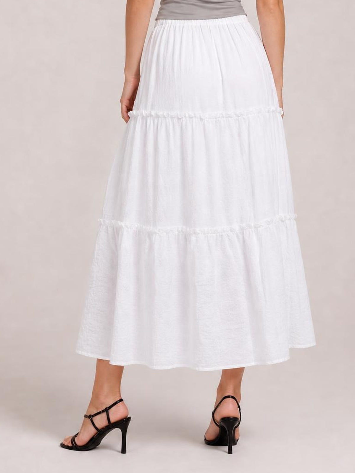Model wears a white frill elastic waist skirt with a lined, tiered maxi silhouette and gathered seams, shown from the back with hands at the sides.