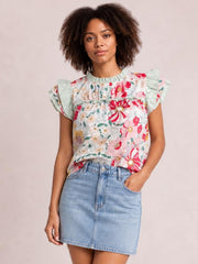 Model wearing an opaque white floral print blouse with red and pink flowers, pleated neckline, ruffle cap sleeves, and a relaxed straight silhouette, posing with one hand in her pocket.