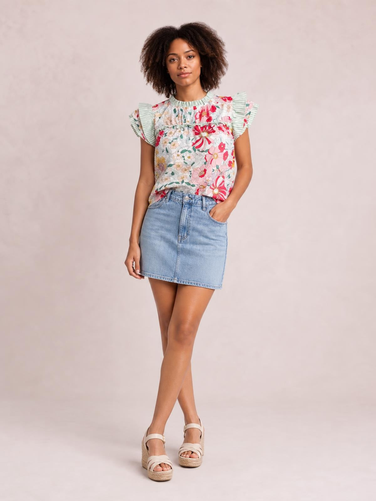 Model poses in a floral print blouse with ruffle cap sleeves, frill detailing, a high neckline, and a relaxed silhouette tucked into a denim skirt.