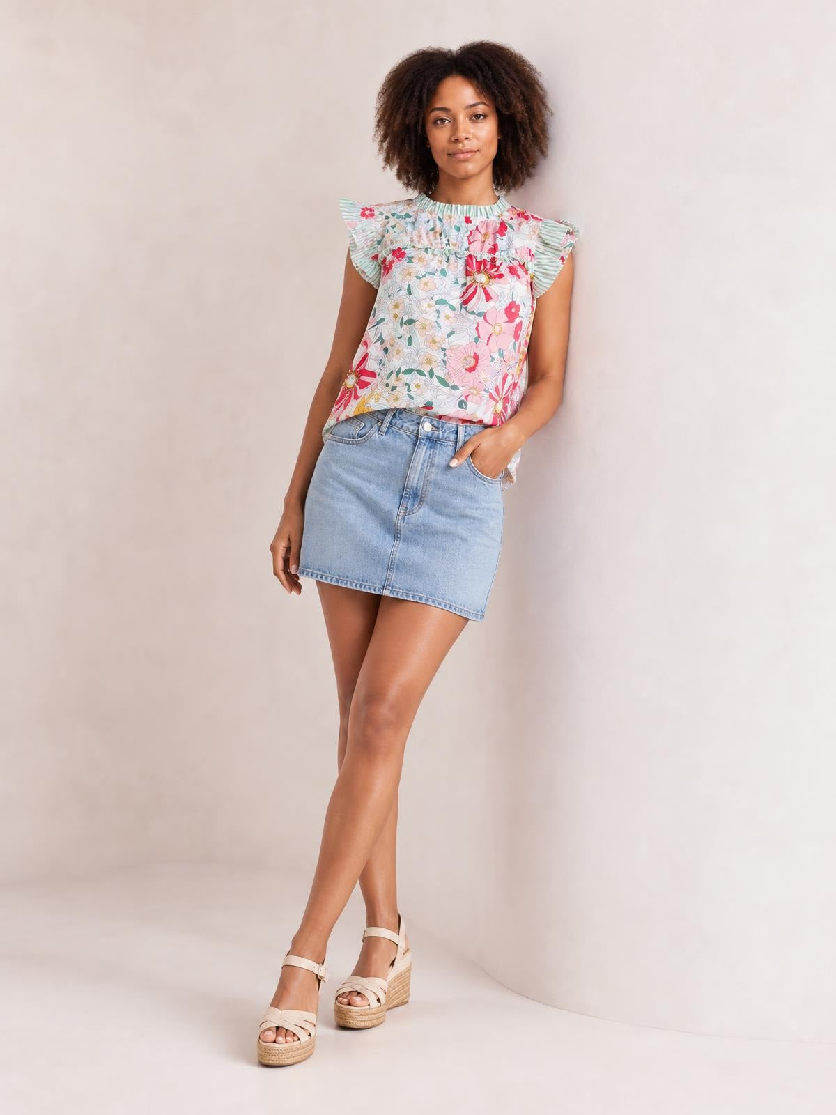 Model wears an opaque floral print blouse in pink, mint, and white with a ruffle-trimmed neckline, flutter cap sleeves, and relaxed silhouette, posing with one hand in her pocket.