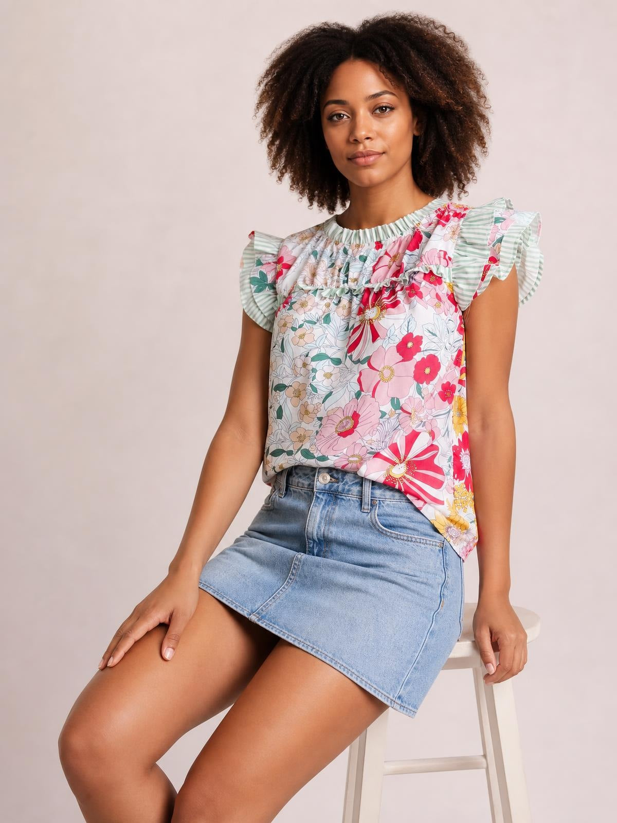 Model seated in a relaxed pose wearing an opaque floral print blouse with ruffle cap sleeves, frill trim, a gathered neckline, and a loose straight silhouette.