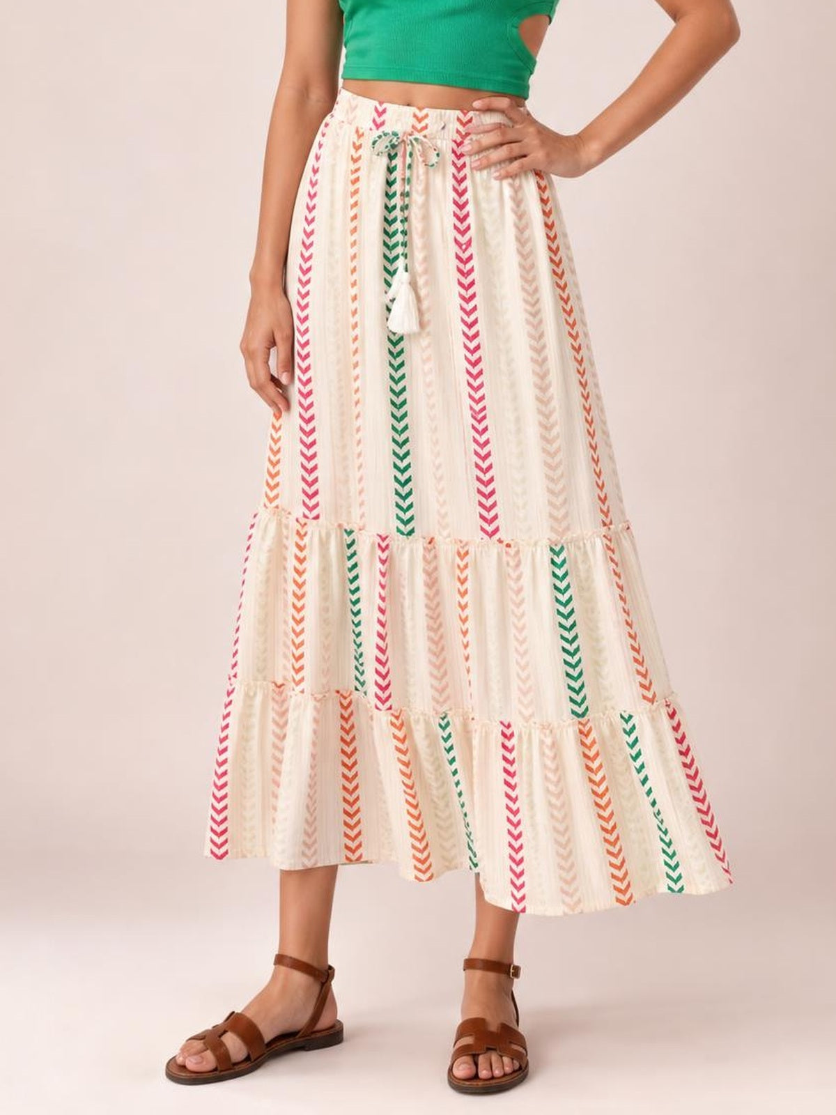 A model wears a flowy cream Frill Printed Maxi Skirt with colorful vertical chevron stripes, a tied waist, tiered frill panels, and an A-line silhouette.
