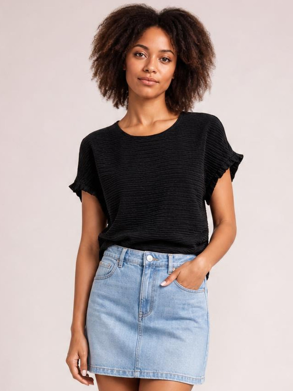 Model wears a black round-neck blouse with frill-trimmed cap sleeves and a relaxed silhouette, posing with one hand in a denim skirt pocket.