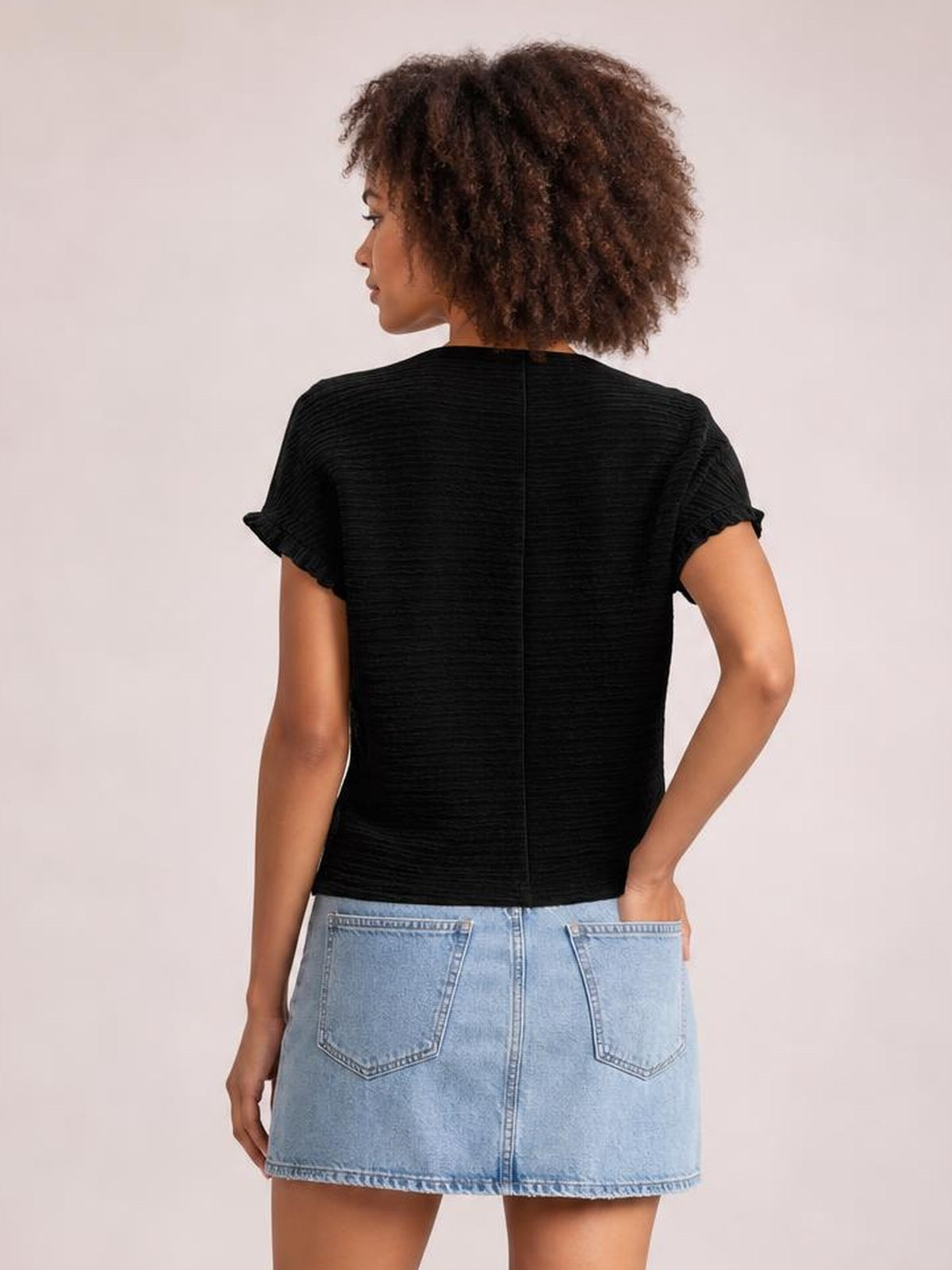 A model poses from the back wearing an opaque black blouse with a round neck, frill-trimmed cap sleeves, and a relaxed straight silhouette.