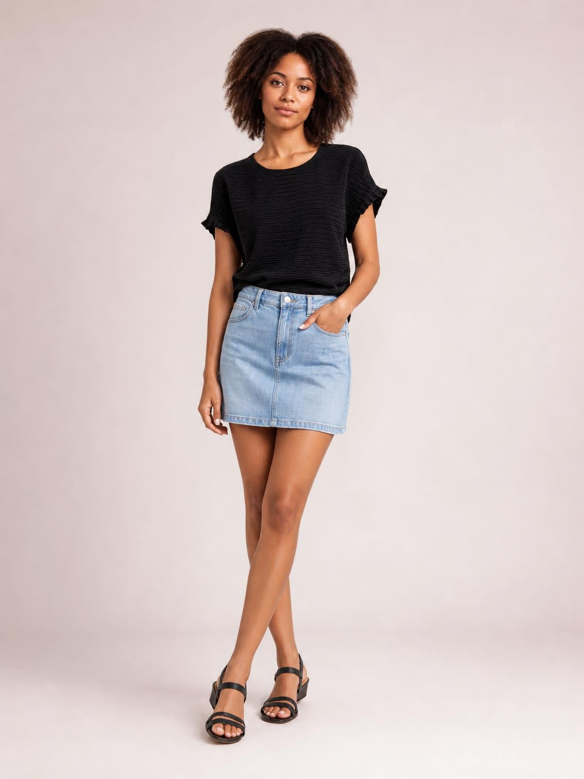 Model wears a black opaque blouse with a round neck, relaxed silhouette, textured finish, and frilled cap sleeves while standing with crossed legs.
