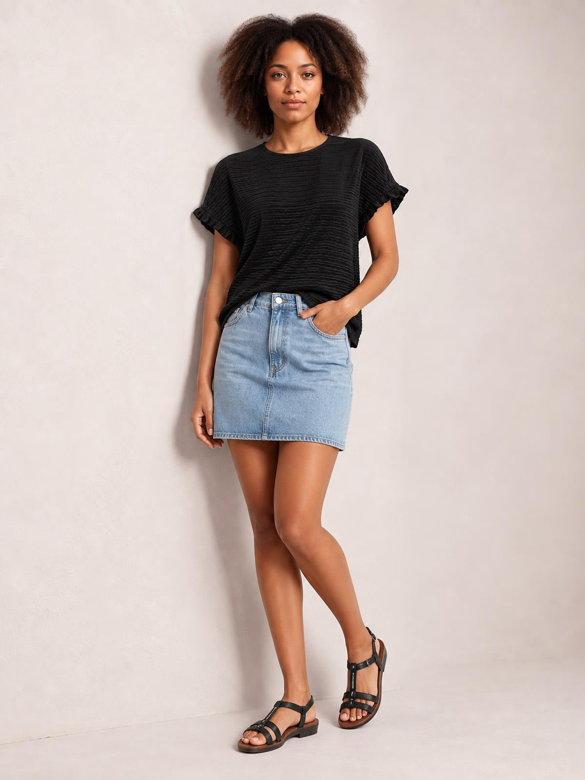 Model wears a black round-neck blouse with short cap sleeves trimmed in frills, a relaxed loose silhouette, and subtle textured construction while posing with one hand in her pocket.