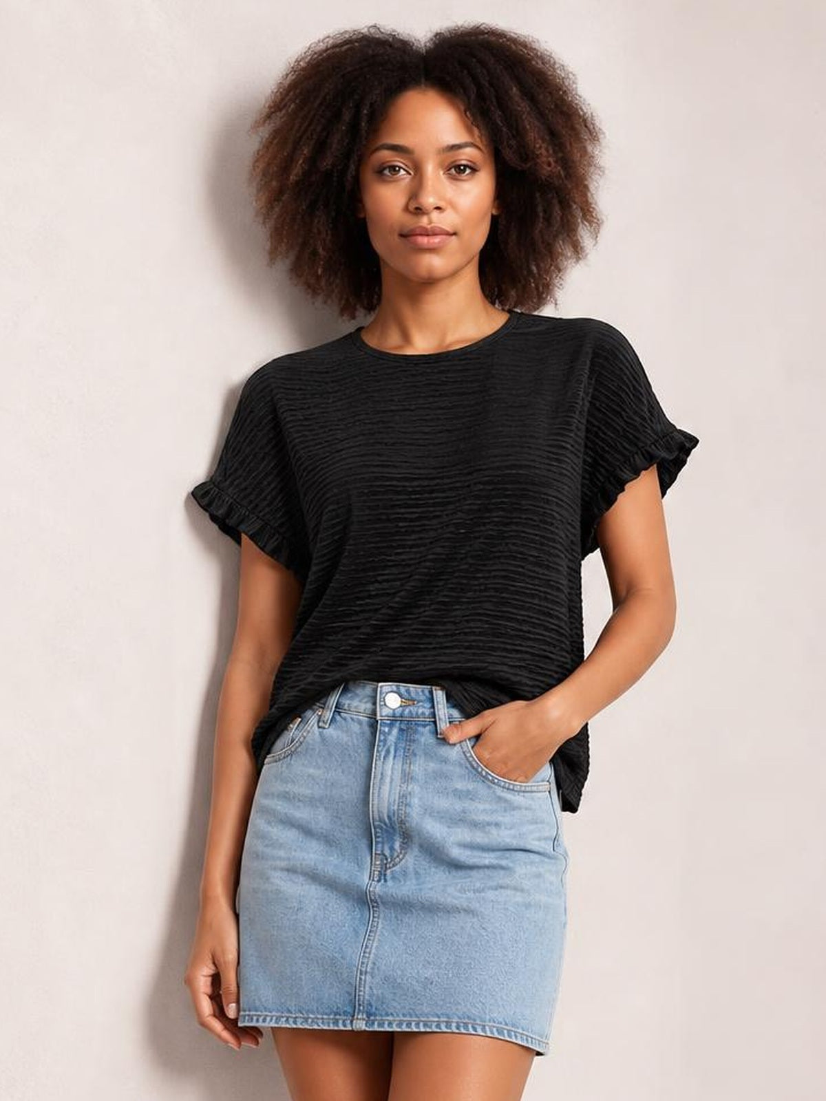 A model wears a black opaque frill round-neck cap-sleeve blouse with a relaxed silhouette and ruffled sleeve trim, posing with one hand in her pocket.