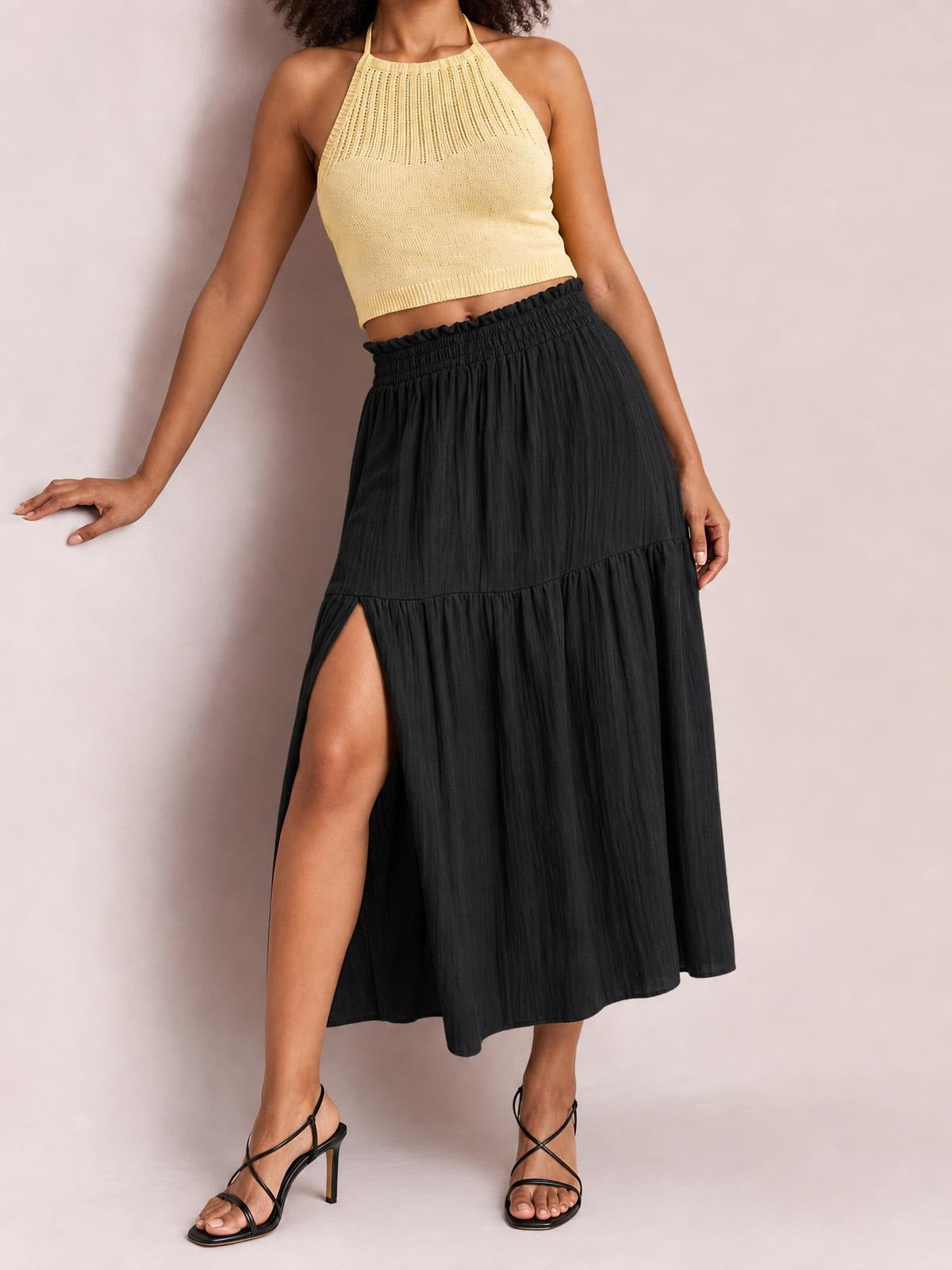 A model poses in a black Frill Slit Skirt with a gathered elastic waist, flowing tiered silhouette, ruffled lower panel, and high side slit.