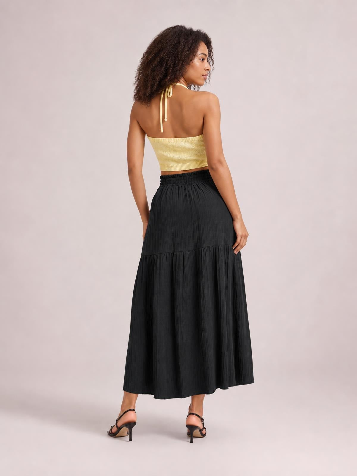 A model stands turned away in a black frill skirt with an elastic waistband, gathered tiered construction, and a flowing ankle-length silhouette.
