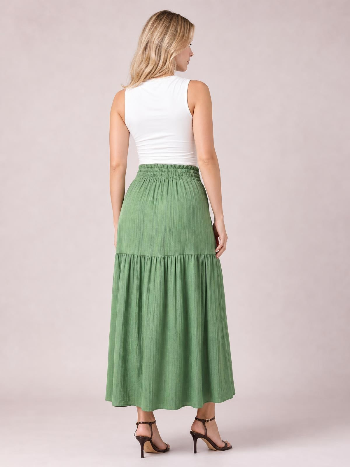 Back view of a model wearing a sage-green frill skirt with a smocked elastic waistband, flowing maxi silhouette, gathered tier, and softly ruffled hem.