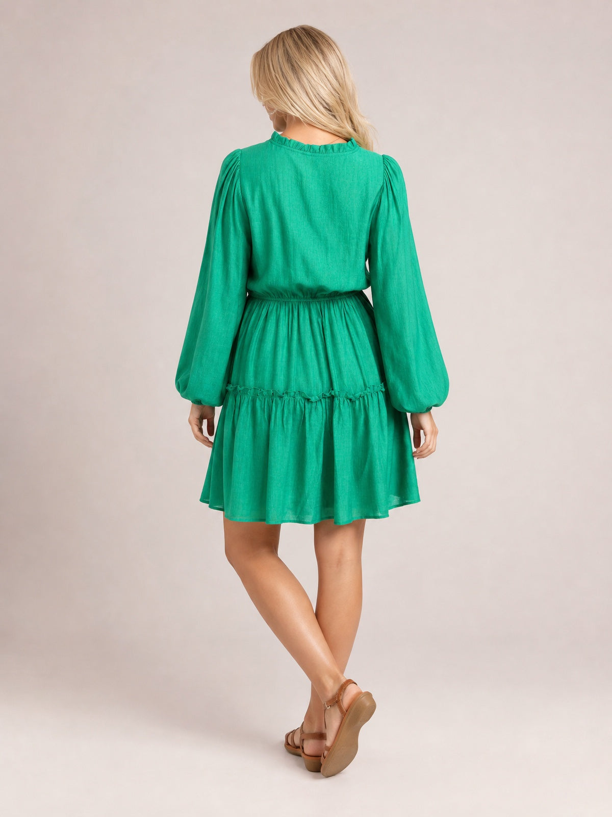 Back view of a model in a turquoise frill mini dress with long gathered sleeves, a defined waist tie, and a flowing tiered skirt, posed with crossed legs.
