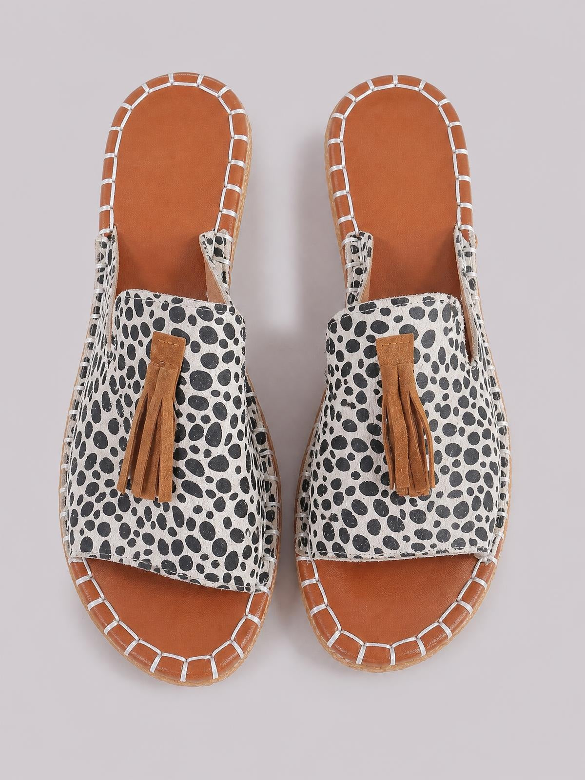 Flat open-toe sandals with a black-and-white leopard-print upper, tan sole, stitched edging, and tan fringe detailing across the vamp.