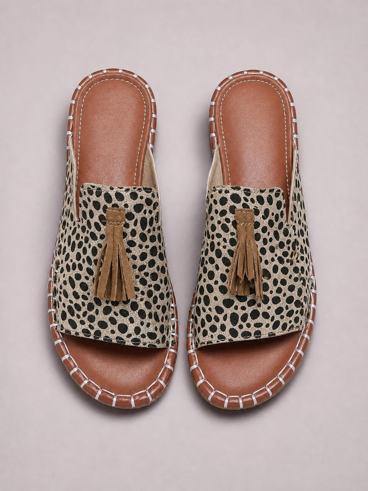Pair of flat open-toe sandals with beige and black leopard print uppers, brown soles, stitched edging, and decorative tan fringe.
