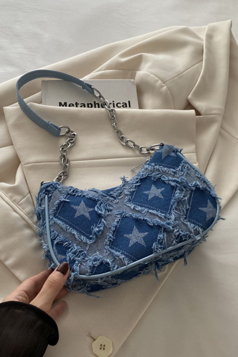 A medium blue denim shoulder bag features a softly curved silhouette, star print, raw fringe trim, and a mixed denim-and-chain strap held by a hand.