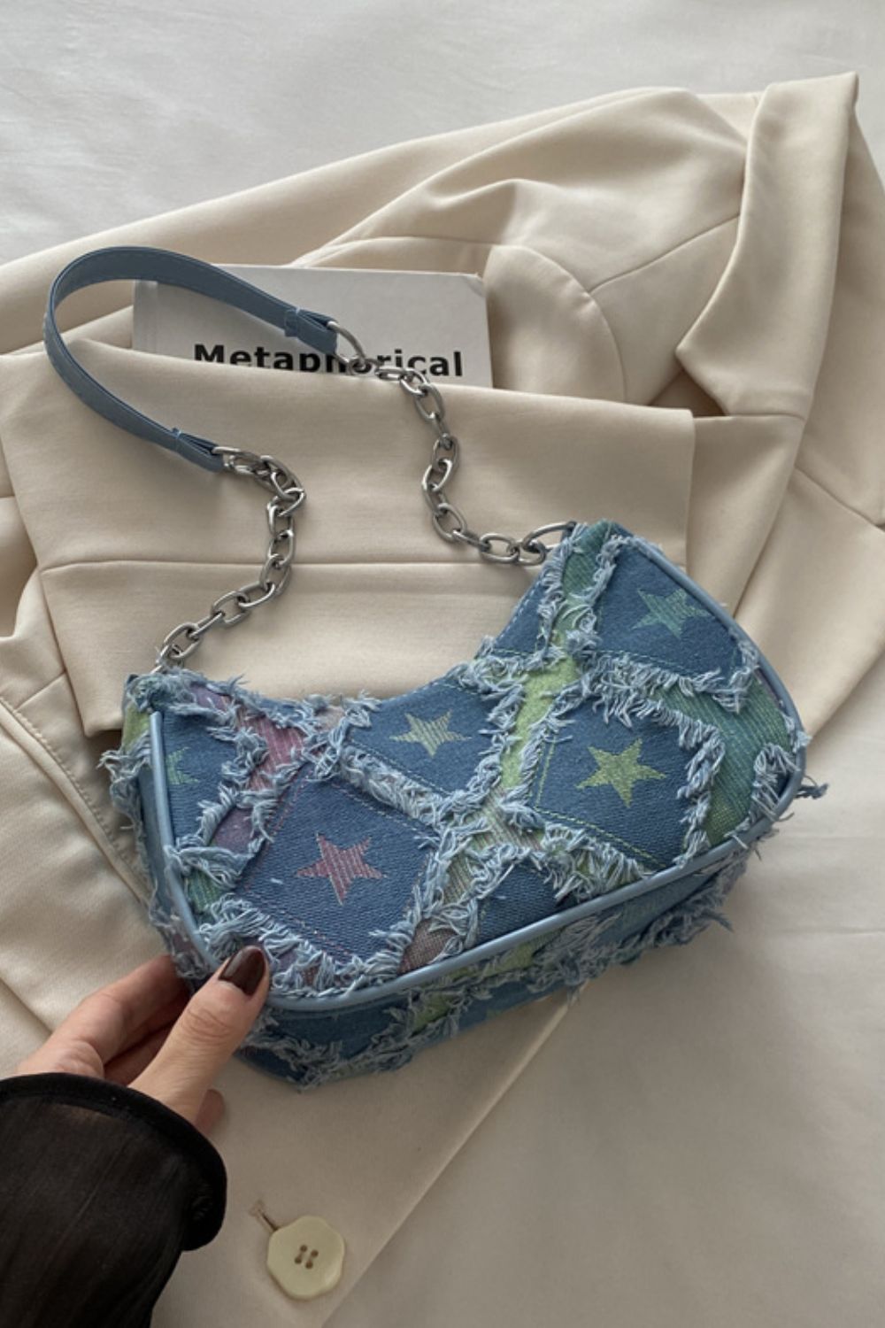 A medium denim shoulder bag features a softly curved silhouette, colorful star print, frayed fringe detailing, and a mixed strap with a silver chain accent.