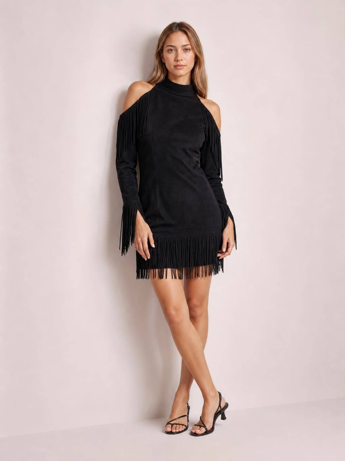 A model poses with crossed legs in a black cold-shoulder mini dress featuring a fitted silhouette, long sleeves, high neckline, and fringe trim at the shoulders, cuffs, and hem.